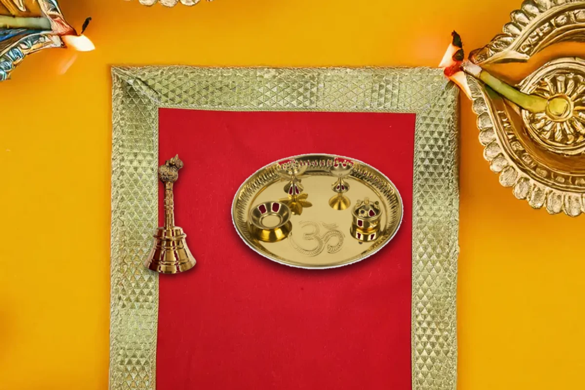 An authentic altar arrangement featuring a brass puja thali set, nandi bell, and lighted brass lamps on a sacred red aasan.